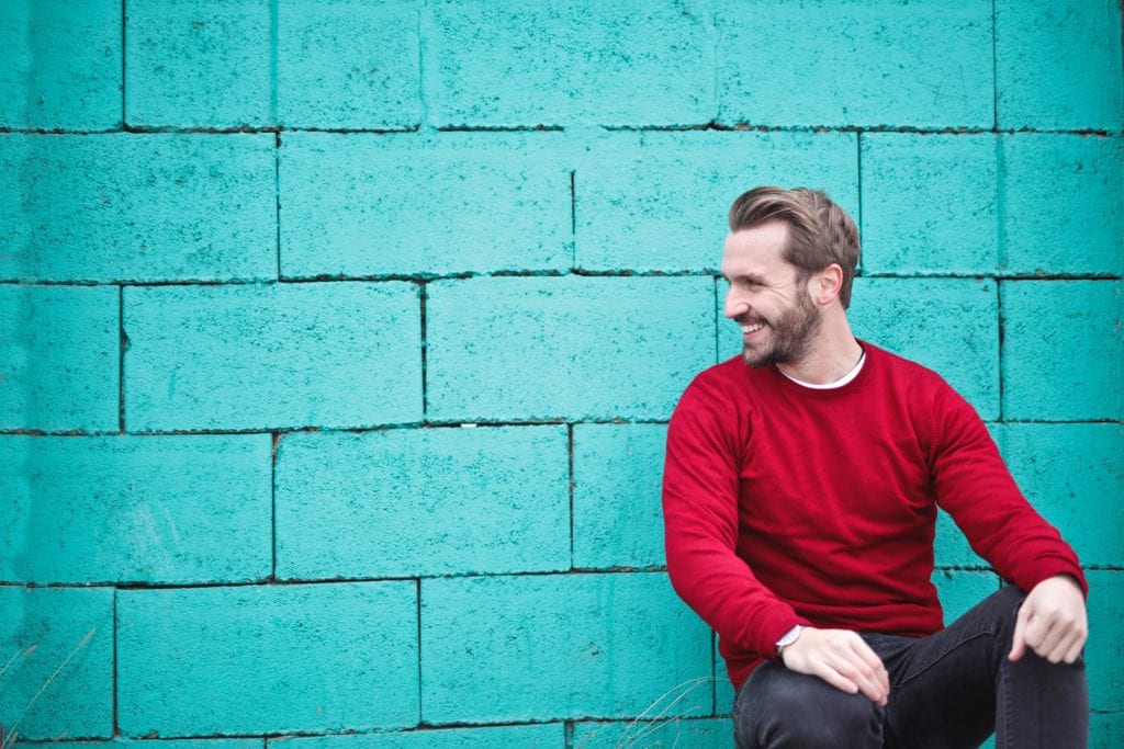 Man wearing red sweatshirt and black pants leaning against a blue wall